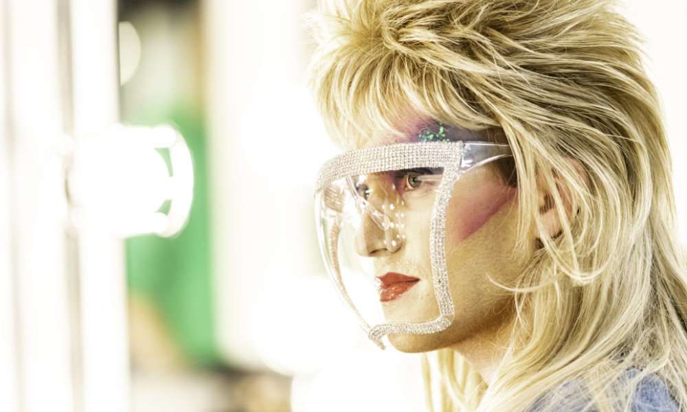 Closeup of student with glam rock hair and makeup wearing a clear face mask.