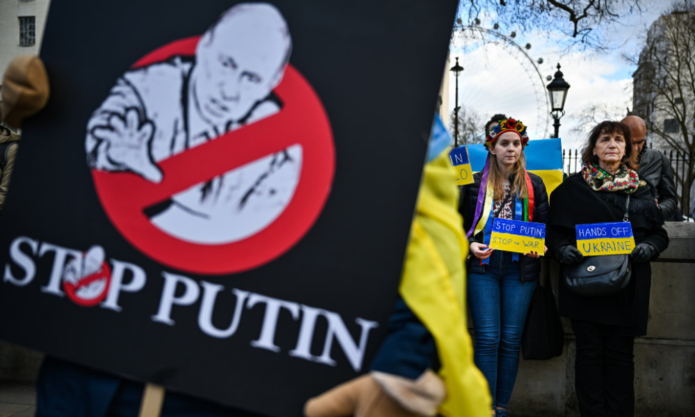 Close-up of anti-Putin rally sign with people in the background holding signs with the Ukraine flag's colors.