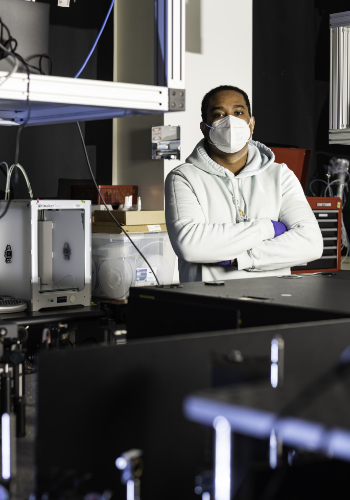 Optics graduate student in a masks poses with arms folded in a lab.