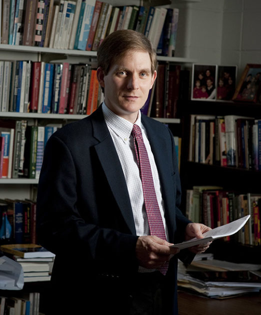 Randall Stone in suit and tie standing in office with bookshelves in background.