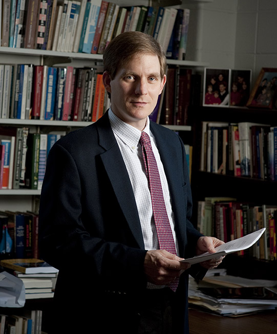Randall Stone in suit in tie standing in office with bookshelves in background.