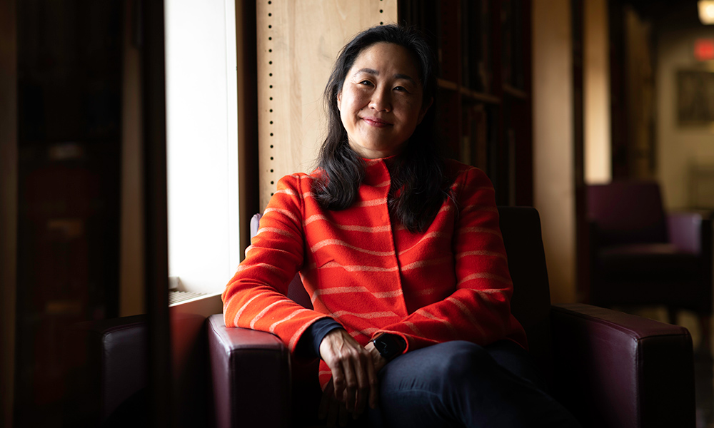 Hwang in red sweater, seated in lounge chair, smiling.