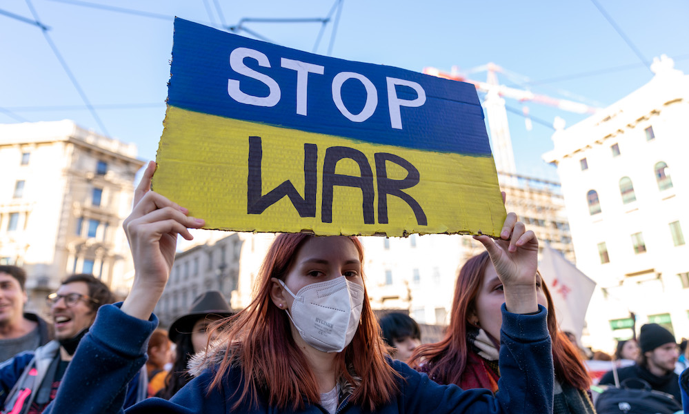Protester in a mask holds up a sign painting as the Ukrainian flag with the message STOP WAR.
