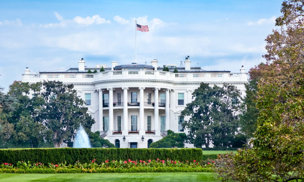 White House with flag atop, blue skies with white clouds, and lush greenery surrounding.