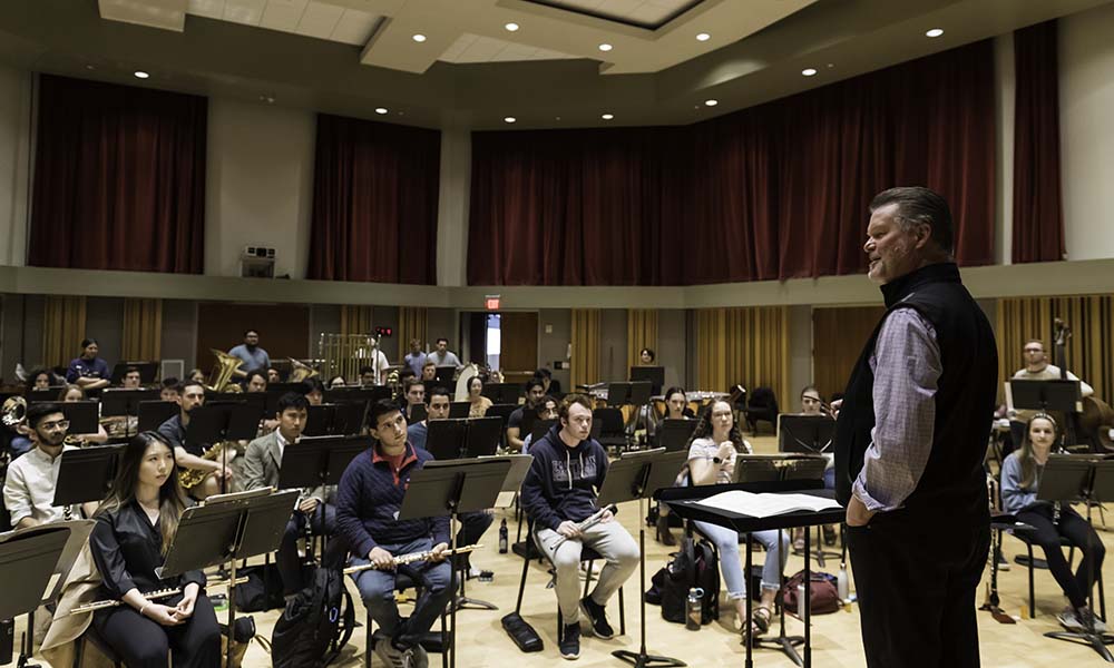 Eastman Wind Ensemble conductor, smiling, talking to musicians who are listening intently during rehearsal.