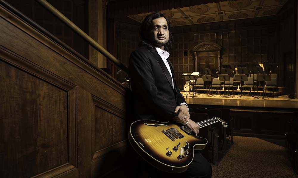 Feature image Eshaan Sood, dressed in tuxedo and in performance hall with guitar resting on knee.