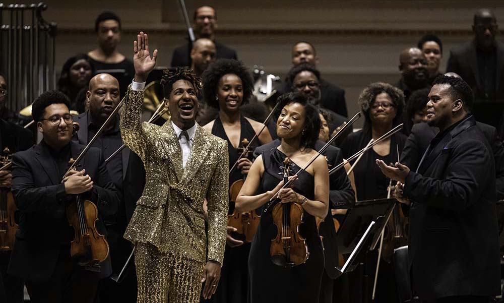Orchestral musicians of African descent standing, clapping on cancer stage with pianist dressed in gold waving.