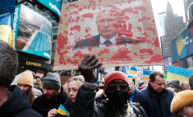 Person in a crowd wears a mask, hat, and sunglasses while holding a sign with Putin's face covered in red handprints.