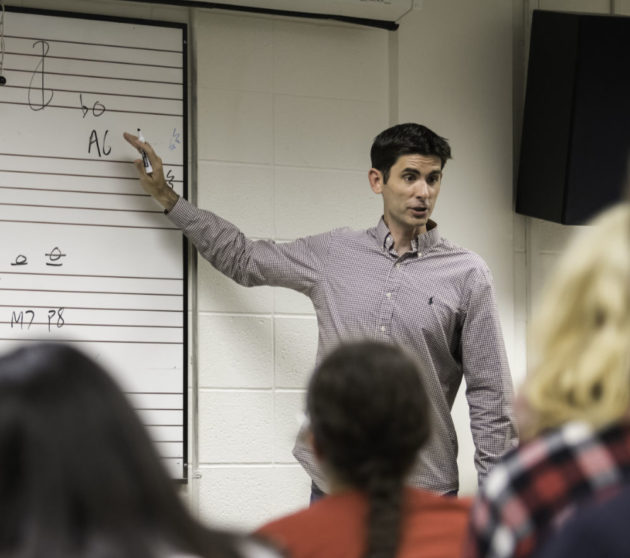 Matt Bailey Shea points at a whiteboard with notations on it while students look on from their seats in class.
