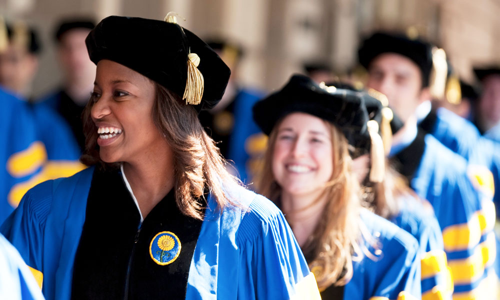 Smiling University of Rochester medical school graduates in caps and gowns lined up outside Eastman Theatre.