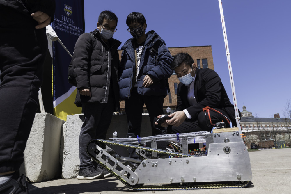 Three students in masks and outdoors stand near an autonomous robot they designed.