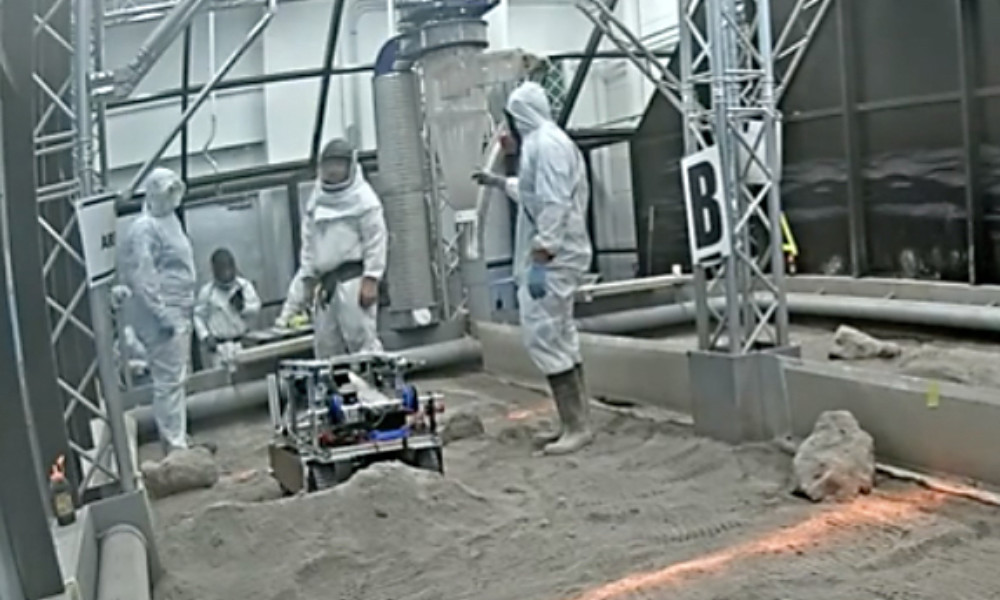 Screenshot of three people in protective gear and a robot on a replica lunar surface.
