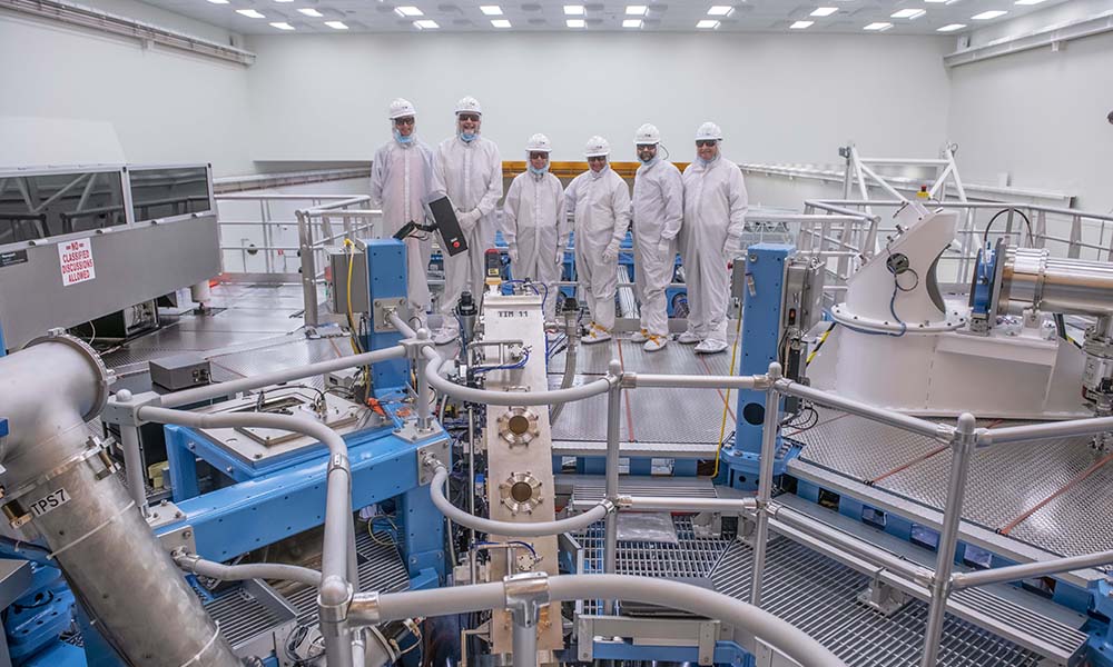 Feature image Six people in body suits and helmets standing in row on deck inside laser lab.