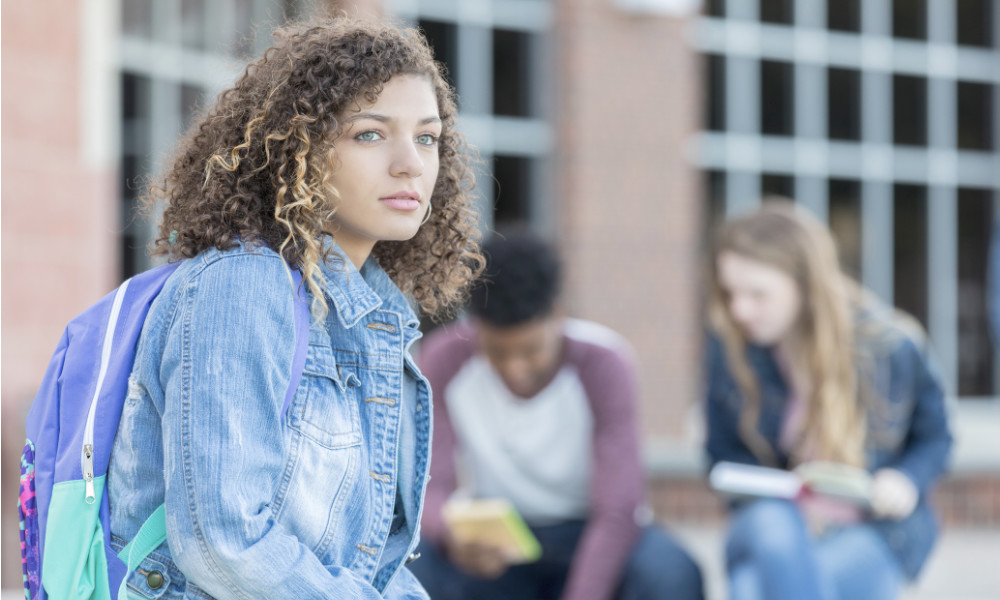 one teen sits alone looking off into the distance as a group of teens sit and talk behind her.