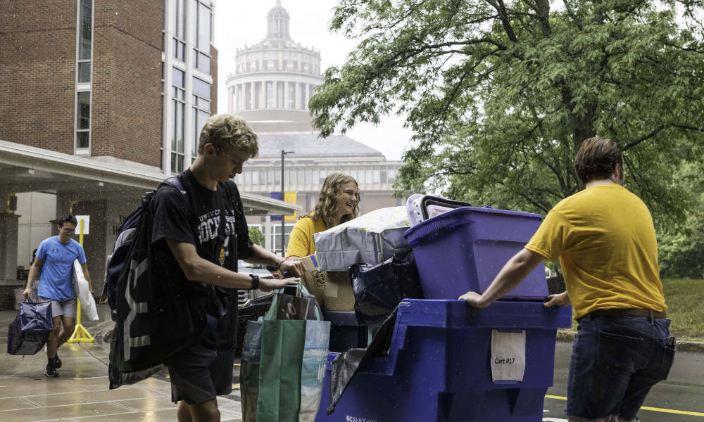 Three people moving into residence halls with luggage atop dollies.