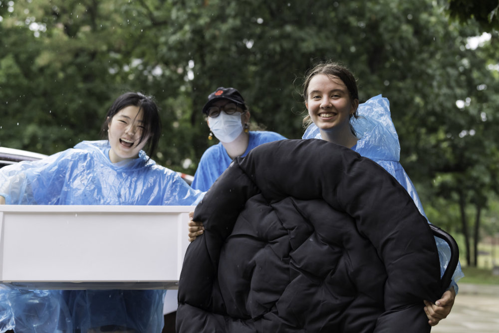 Three students in blue rain ponchos moving luggage and gear.
