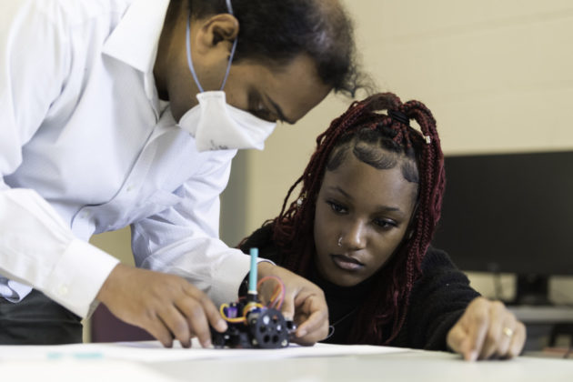 Professor in mask and student lean over small robot.