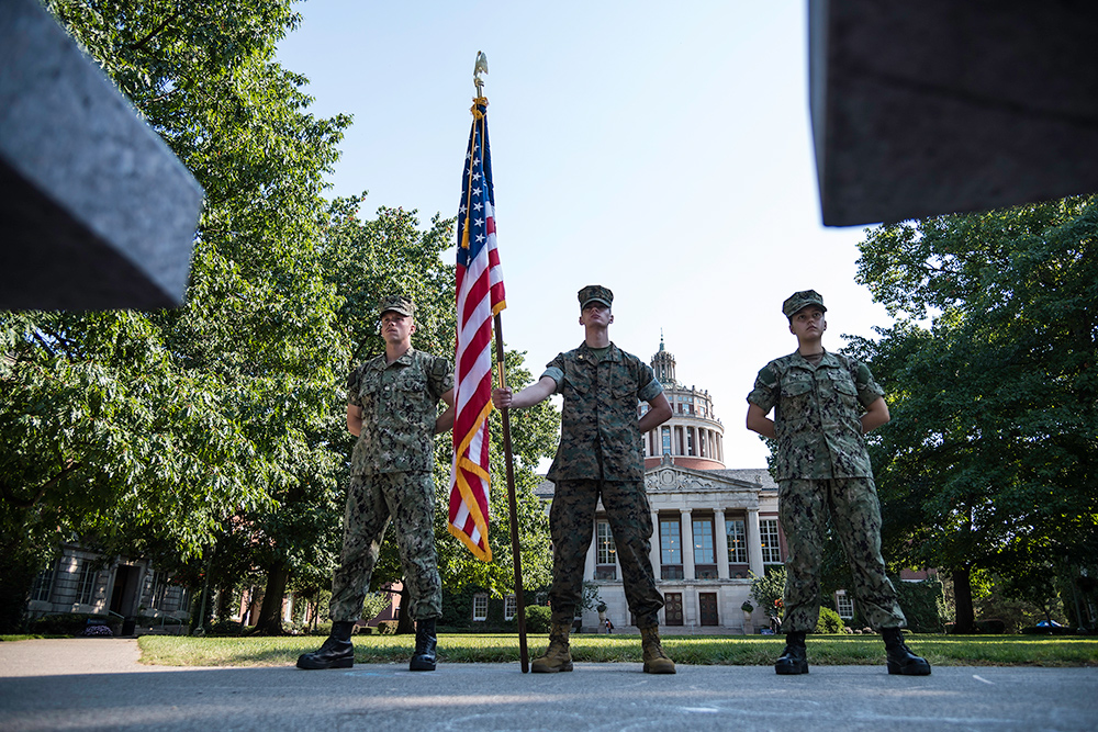 Three students in military fatigues stand at ease in front of Rush Rhees Library on the quad, the student in the middle holding an American flag.