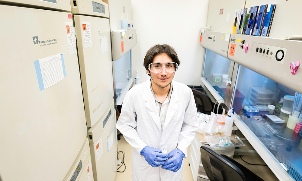 Awad in a white lab coat and blue gloves stands in a lab full of equipment.