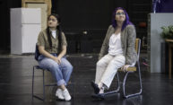 two student actors seated on a stage, both looking away from each other.