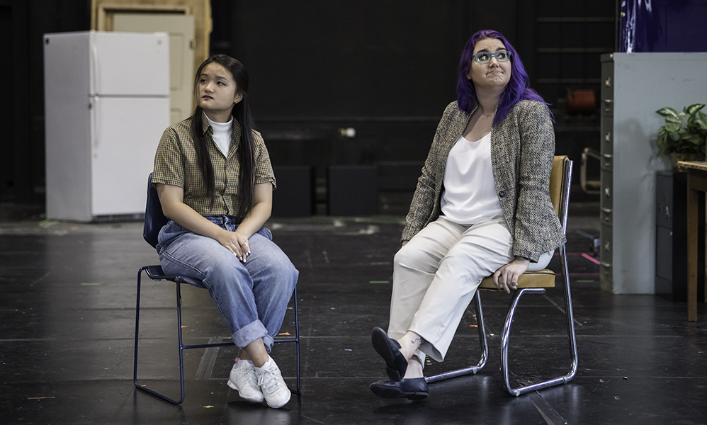 Feature image two student actors seated on a stage, both looking away from each other.