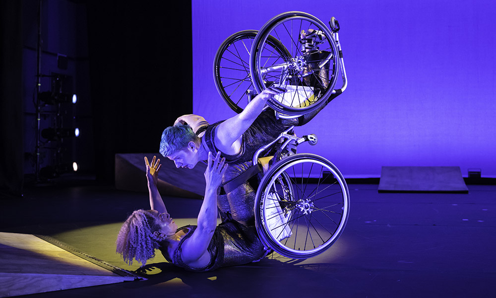 Laurel Lawson hovers above the stage, arms and wheels lifted, supported by Alice Sheppard who partners her from below. Alice is on her back, lifting Laurel, locking eyes and opening her arms in embrace. They are surrounded by small wood ramps and bathed in cool blue light. Laurel is a white dancer with cropped blue hair and Alice is a multiracial Black woman with short curly hair; their skin, shimmery costumes and chairs reflect the light.