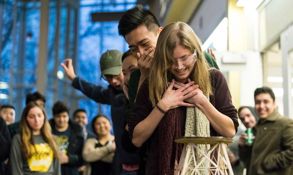 a line of four students, each making different expressions of excitement or laughter, looking at a sculpture made of popsicle sticks as if expecting it to collapse.