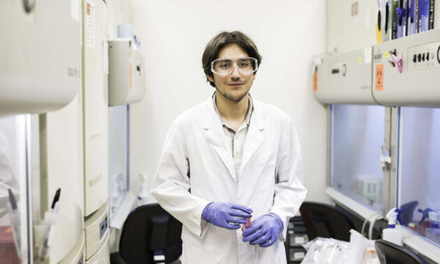 student in a white lab coat and blue gloves stands in a lab full of equipment.