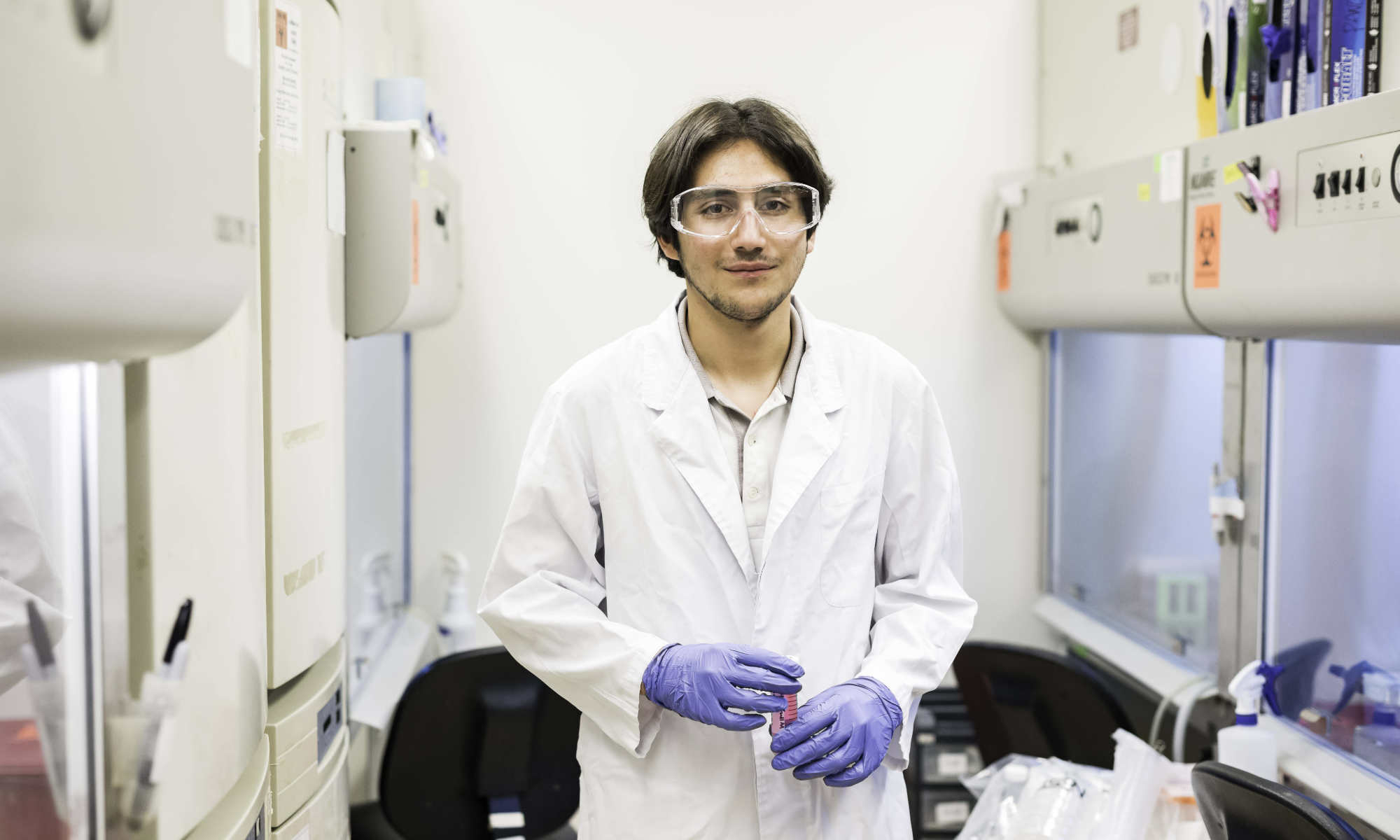 Feature image student in a white lab coat and blue gloves stands in a lab full of equipment.