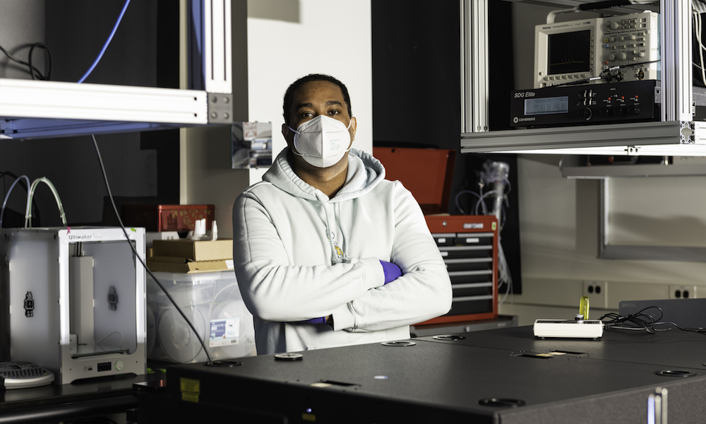 Optics graduate student in a masks poses with arms folded in a lab.