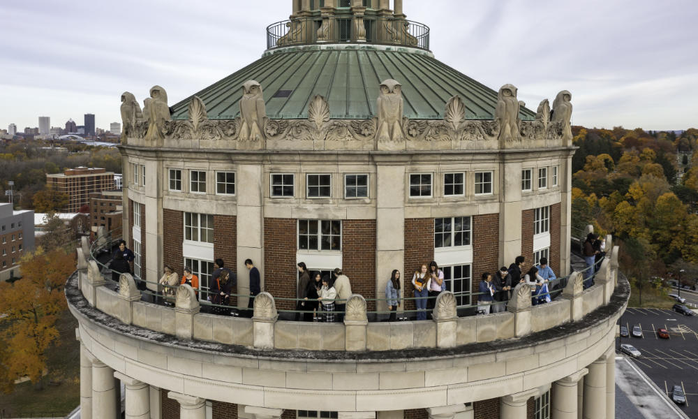 Feature image Drone photo of Rush Rhees Tower with people on tour and the city of Rochester in the background on a cloudy day.