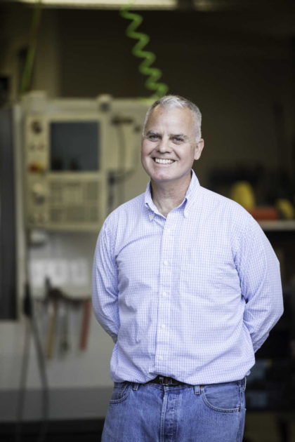 Chris Muir smiles and stands in a fabrication shop with his hands behind his back.