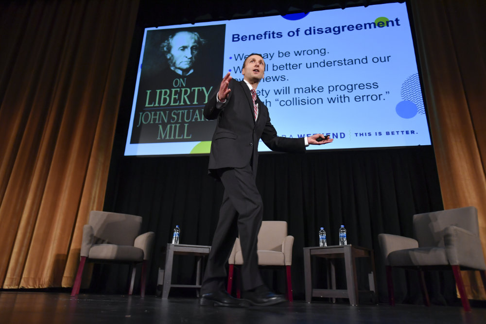 David Primo on stage discussing free speech in front of a PowerPoint screen with the headline "Benefits of disagreement" and a photo of John Stuart Mill's "On Liberty" book cover.