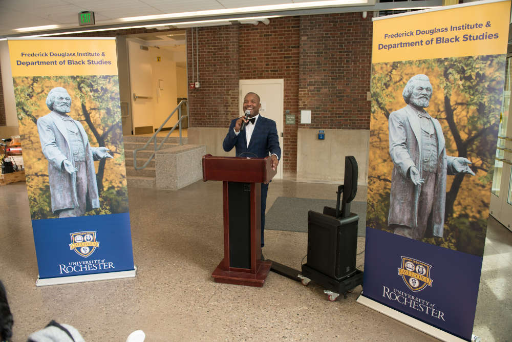 Jeffrey McCune Jr. at a podium between two retractable banners featuring images of a Frederick Douglass statue. 