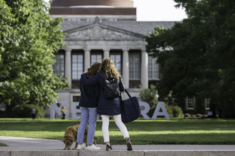 Two people seen from behind give each other a side hug with a dog at their feet on the Eastman Quad. 