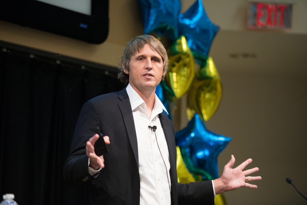 Michael Hasselberg delivering a talk on stage with blue and yellow balloons behind him. 