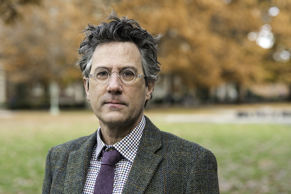 Aaron Hughes in tie and tweed jacket against autumn campus background.