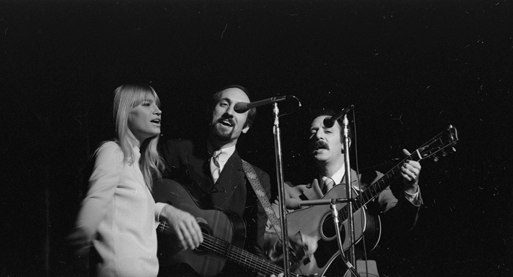 archival photo of Peter, Paul, and Mary singing on stage behind microphones.