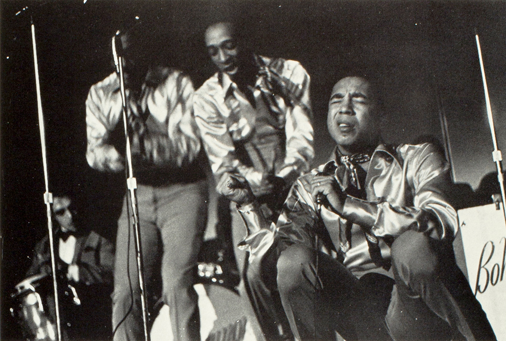 archival concert photo of Smokey Robinson, sitting on the stage and singing.