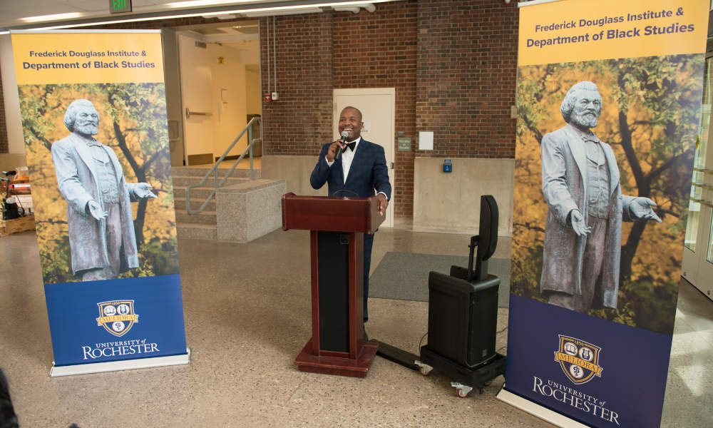Jeffrey McCune with a microphone at a podium between two pop-up banners that say Frederick Douglass Institute & Department of Black Studies.