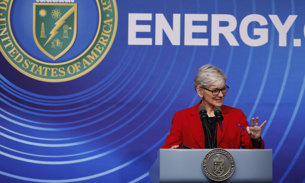 Jennifer Granholm in a red business suit at a podium against a blue background with a US seal and the word ENERGY.