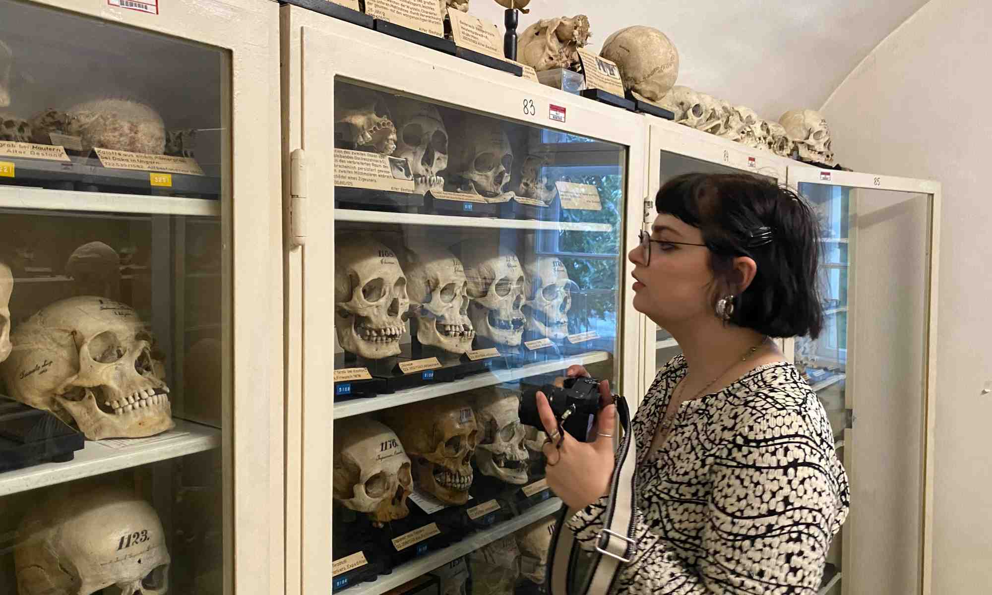 Student holding camera stands in front of a case full of human skulls at a dark tourism destination.