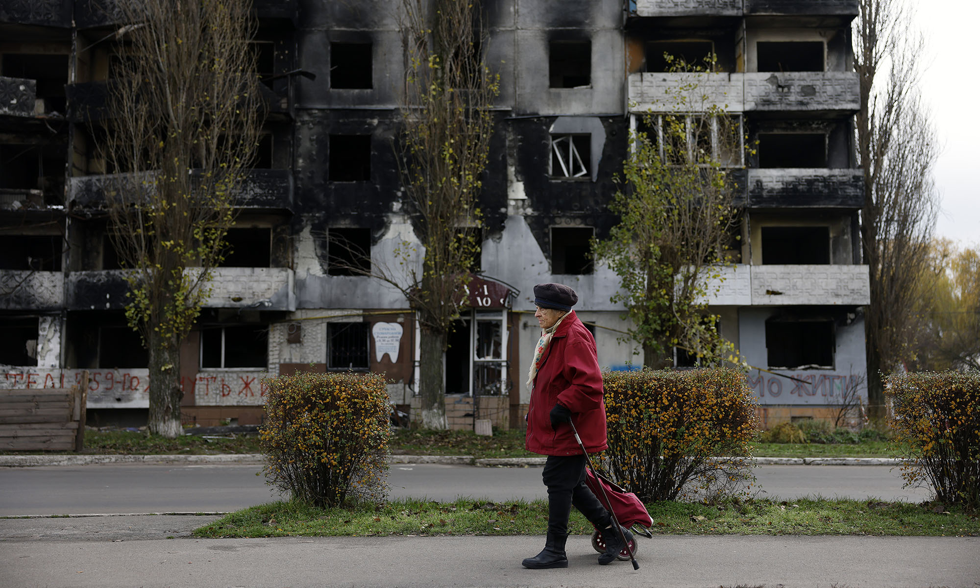 Ukrainian person in hat and coat walks with cane on sidewalk past bombed building to illustrate what is happening in Ukraine.