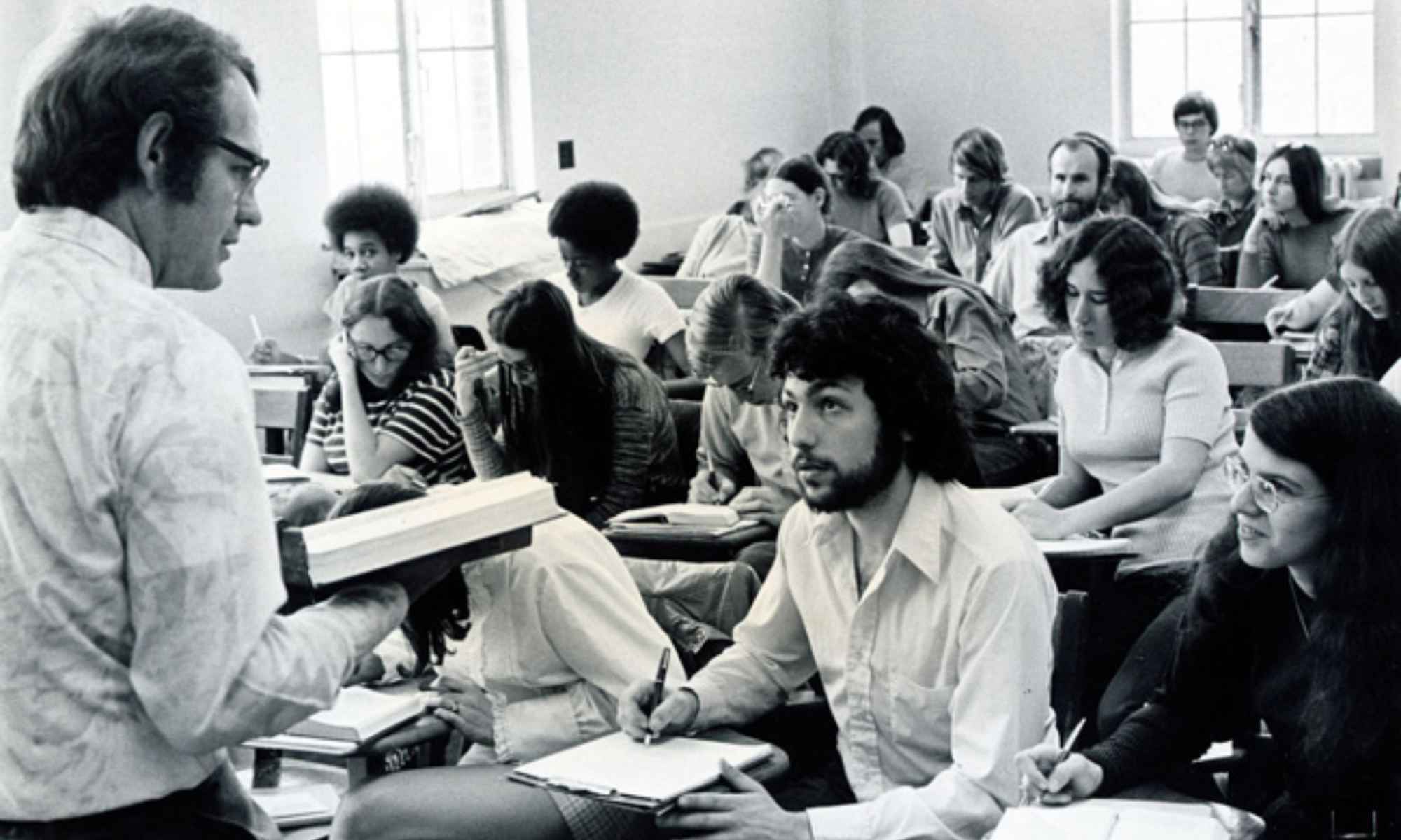 Feature image Black and white archival photo of Russell Peck holding a book and teaching in front of a classroom full of students.