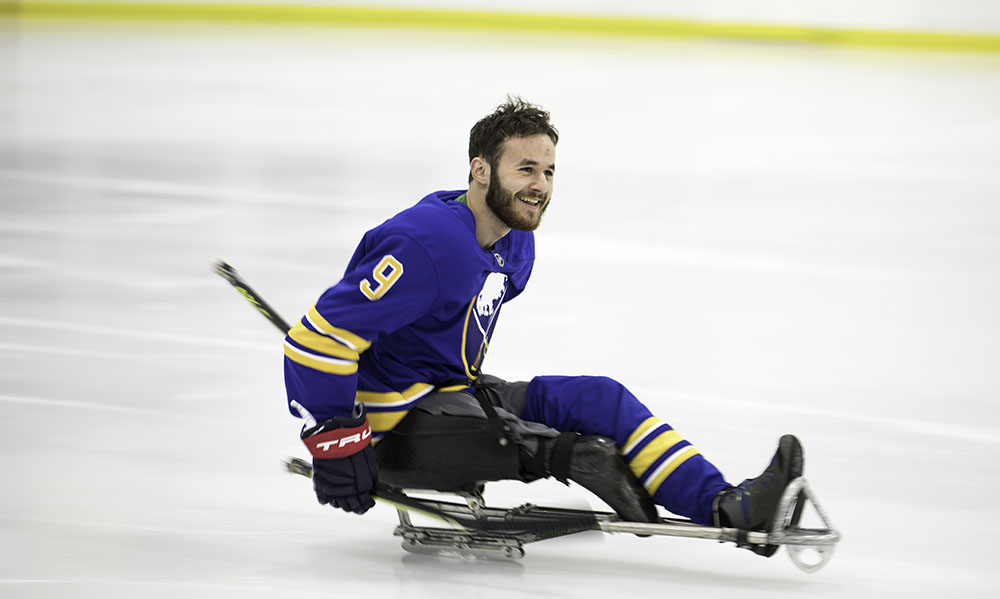 Becker, smiling and gliding across ice rink in Sabres uniform, mounted on hockey sled.
