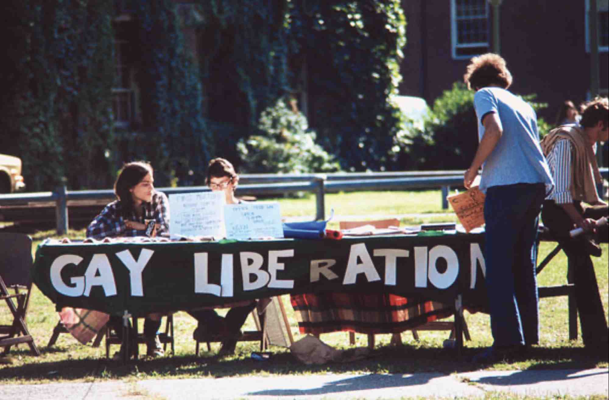 Two people seated at a table outdoors with a table drape that reads "gay liberation front" while a person seen from behind peruses at materials on the table.