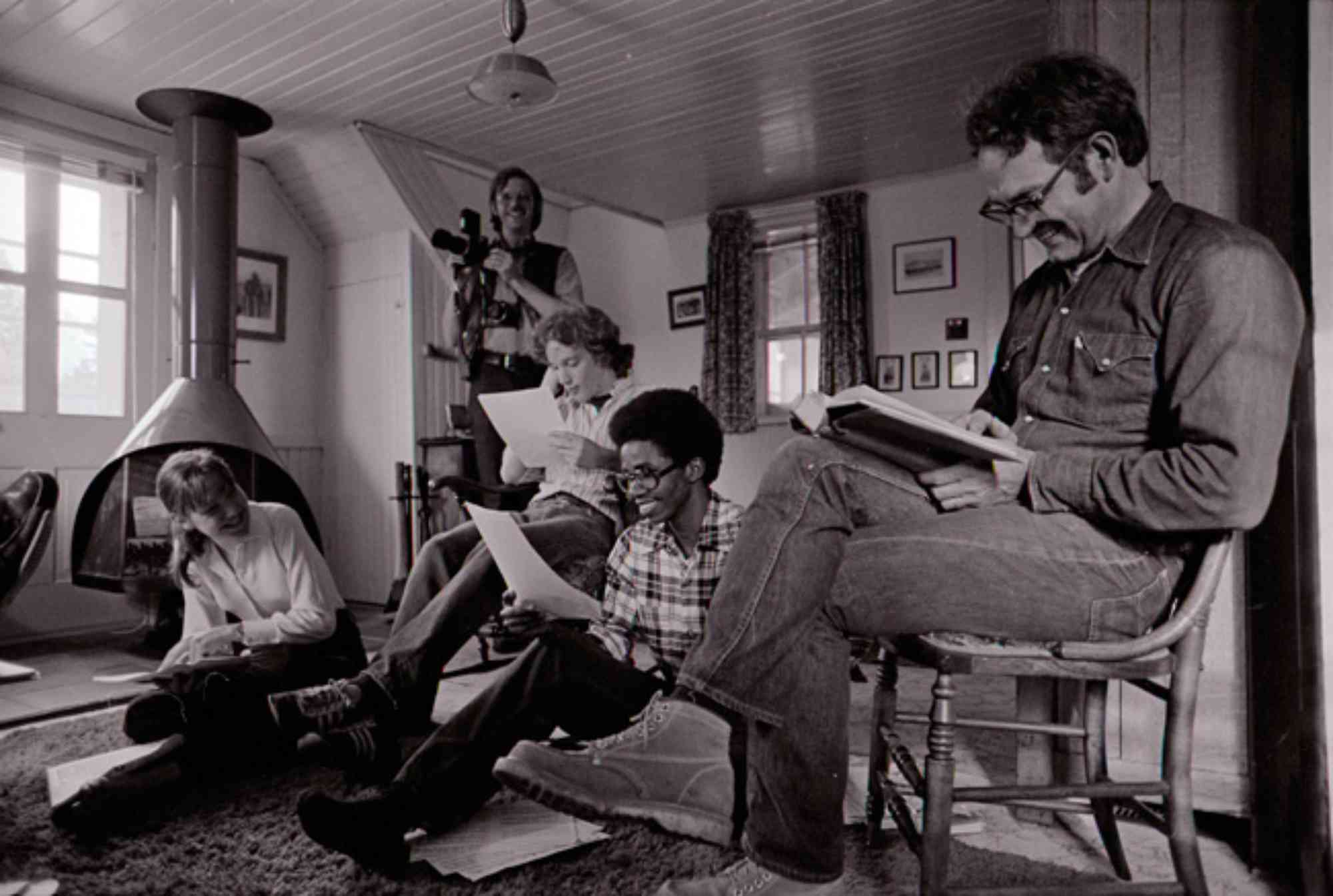 Black and white archival photo of Russell Peck reading indoors at his farm with several students seated nearby.