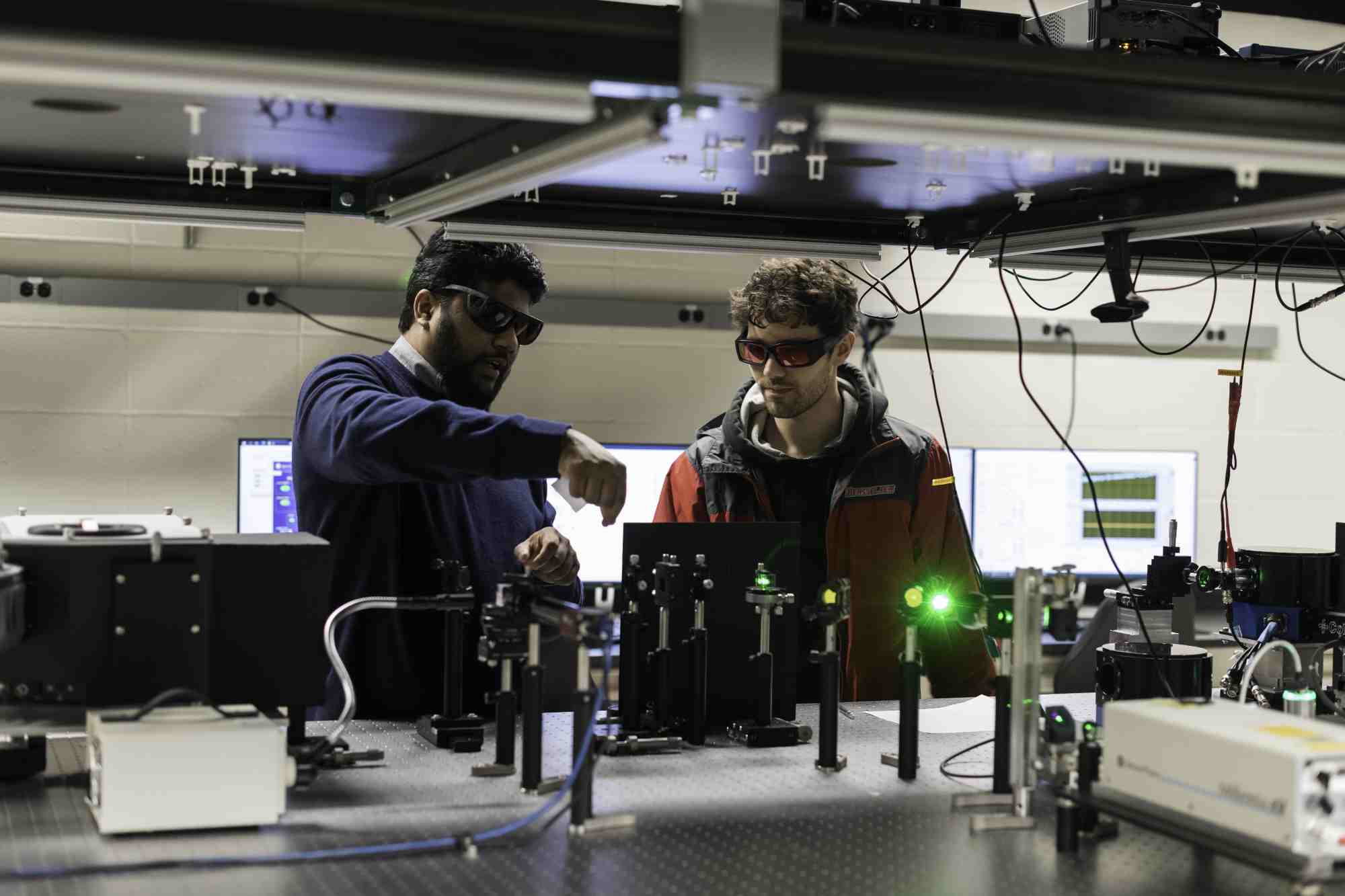 Two people in goggles in an optics lab gesturing over a laser array. 