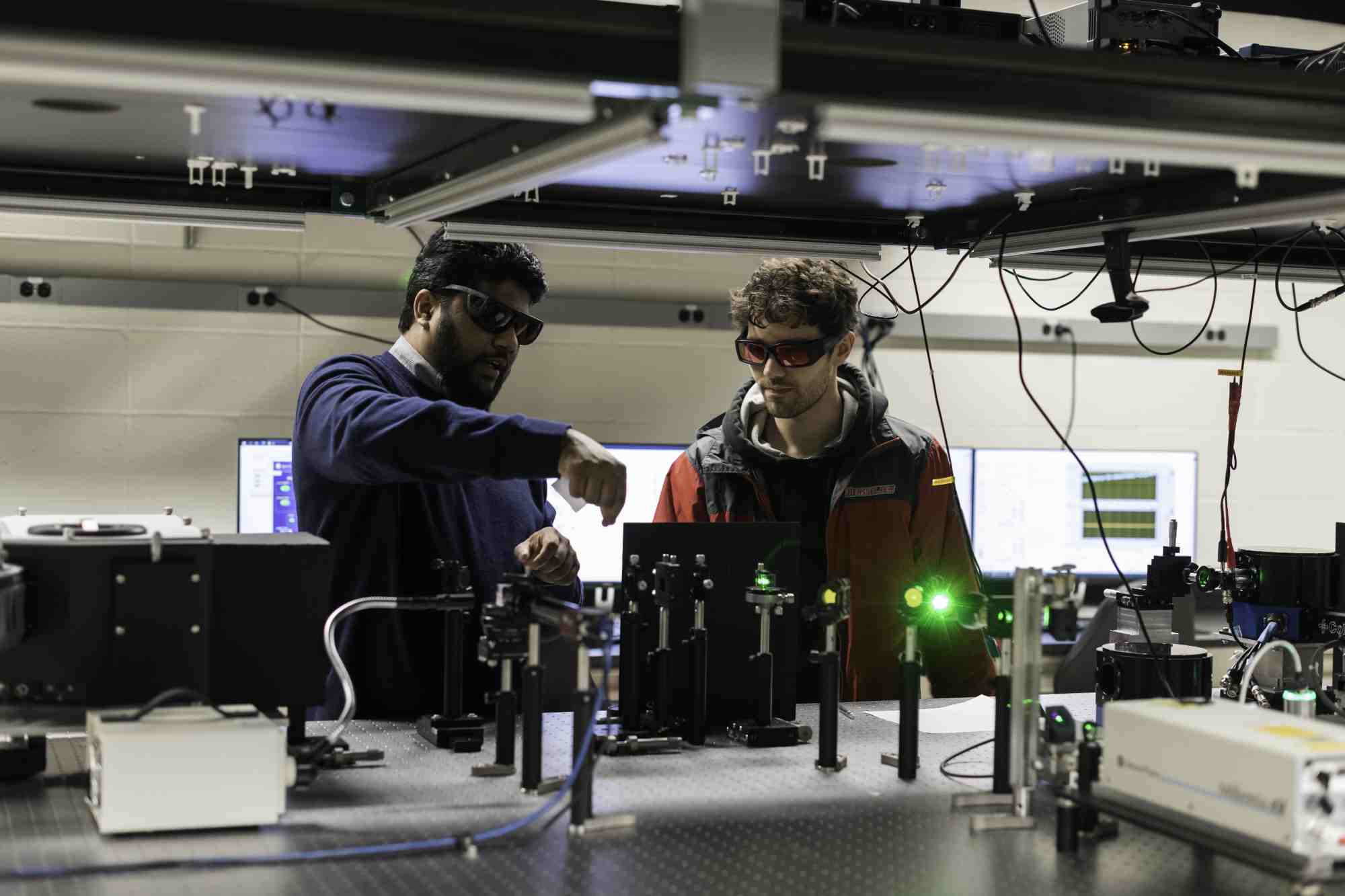 Two people in goggles in an optics lab gesturing over a laser array.