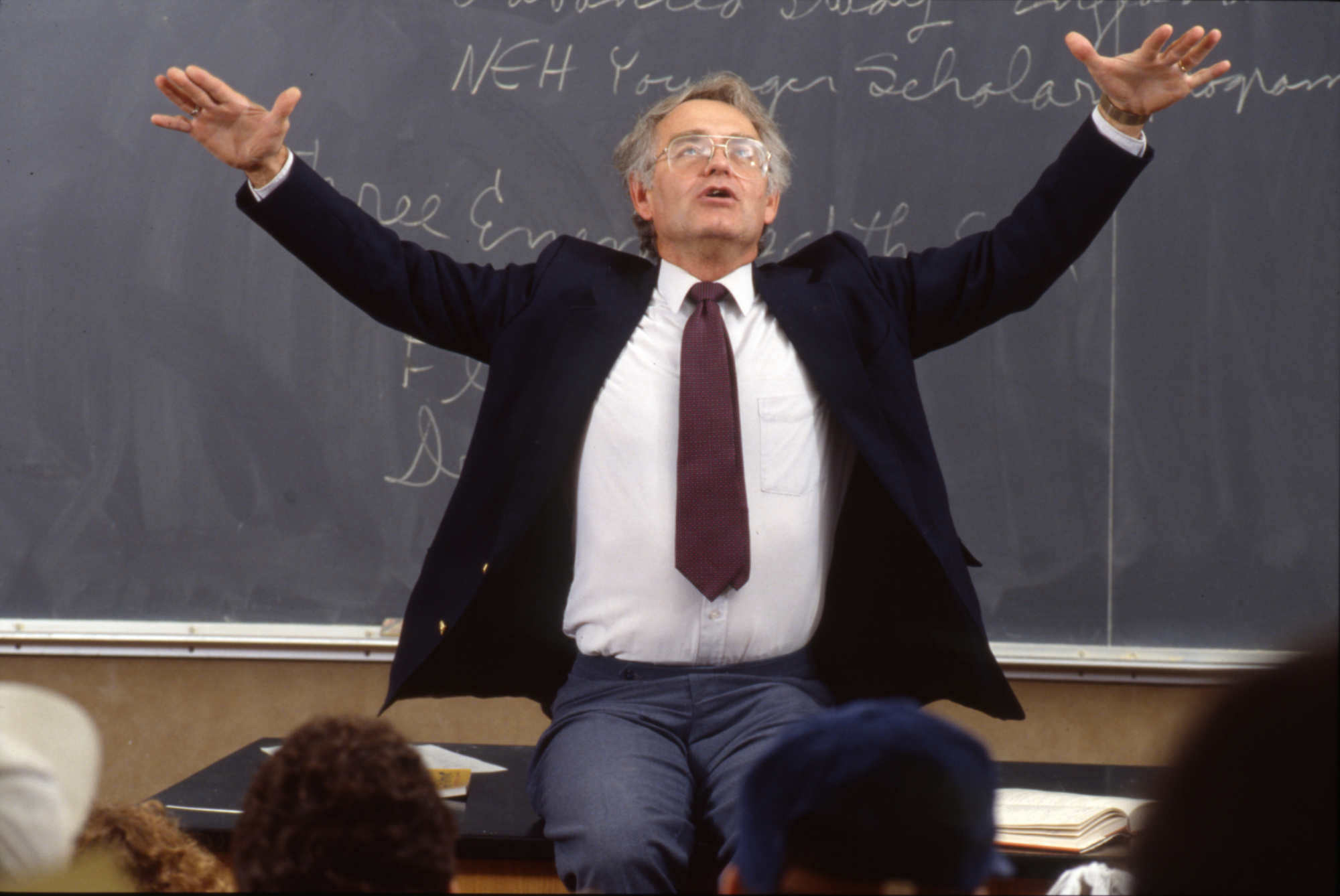 Russell Peck at the front of a class full of students with his arms raised theatrically.